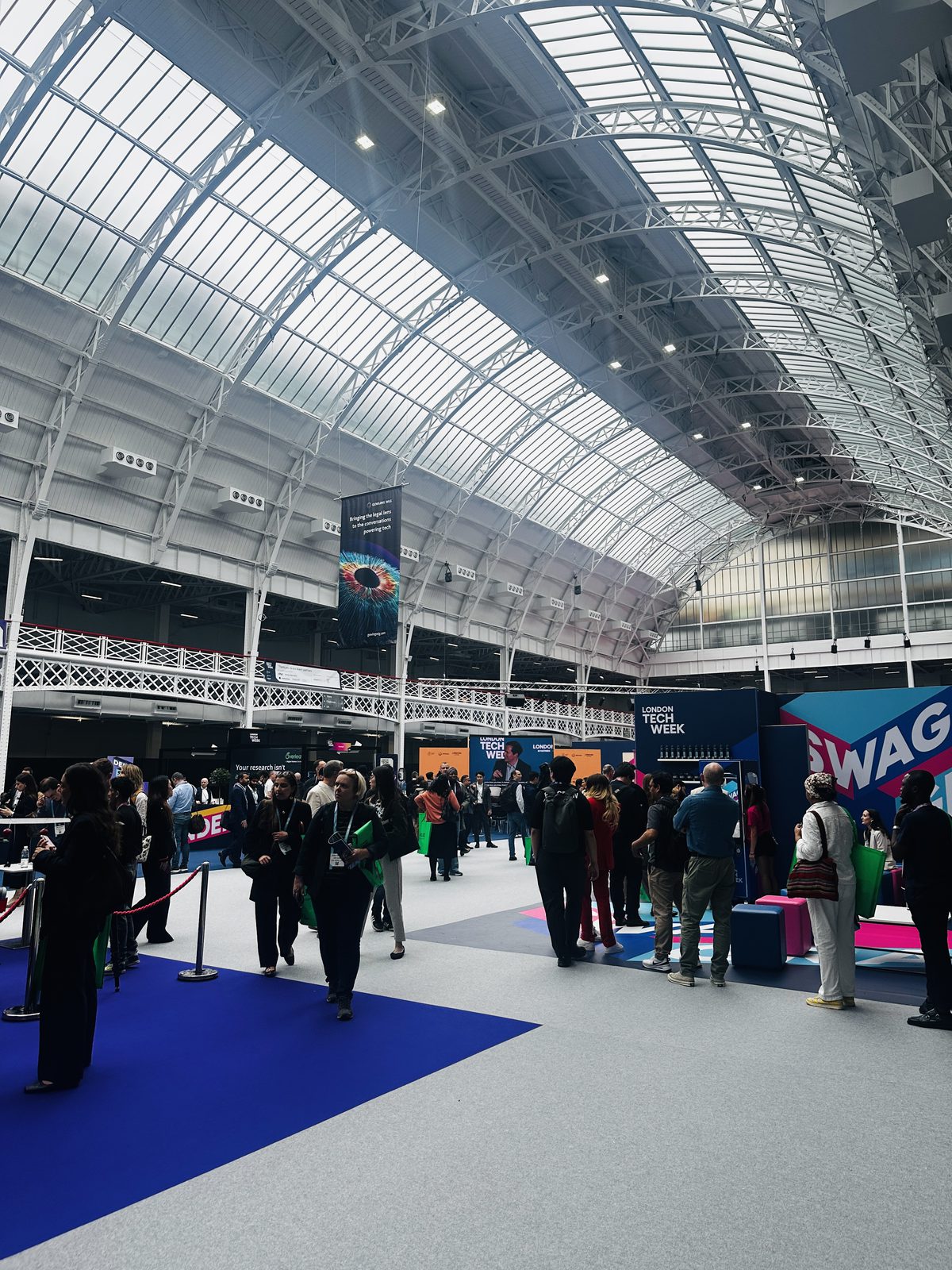 The vast arched glass hall of Olympia London during London Tech Week, crowds and stands below